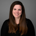 Professional portrait of a smiling woman with long brown hair, wearing a black top, suitable for media representation and branding purposes.