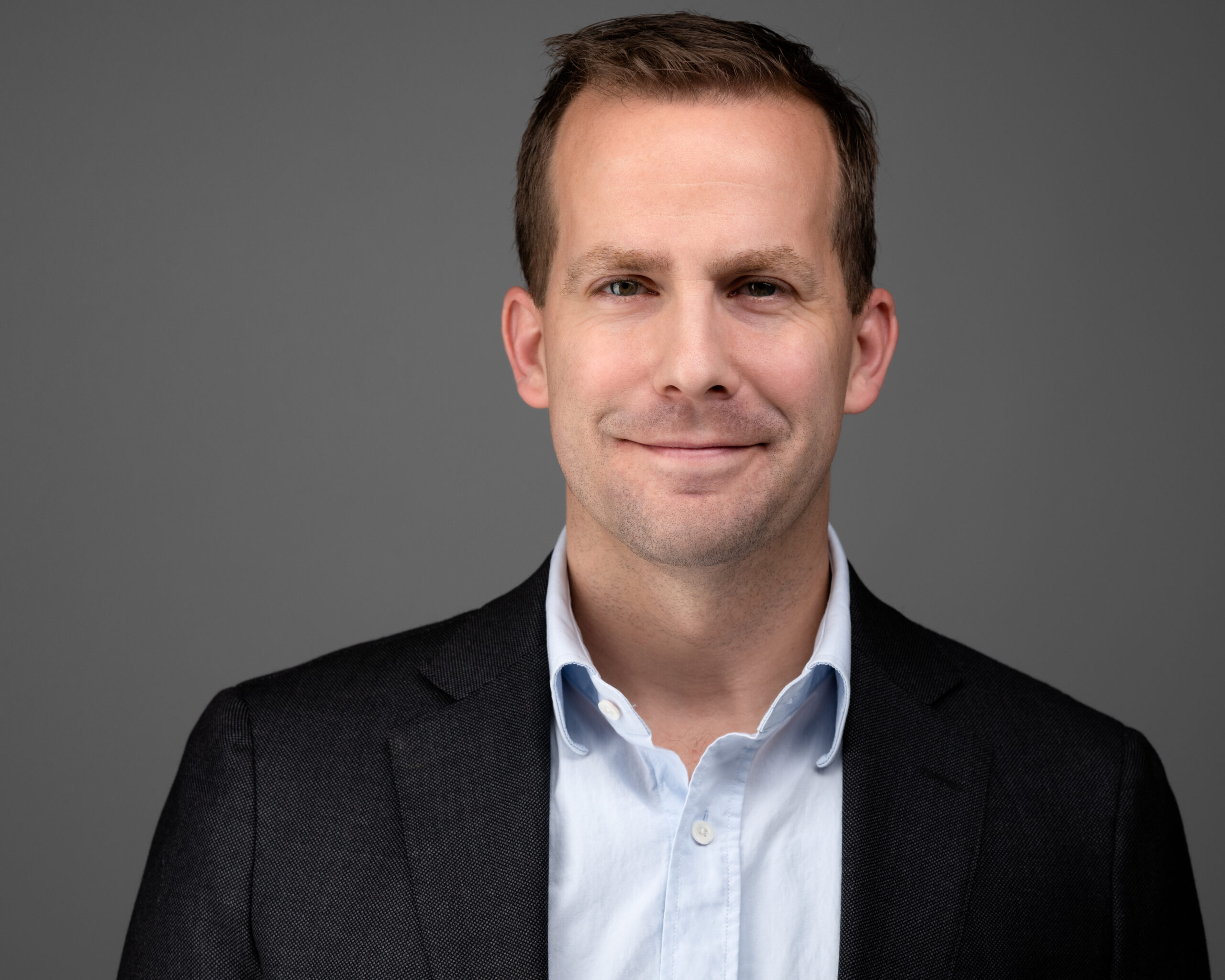 Portrait of Mathew Harkins wearing a blazer and dress shirt, centered on a gray background.